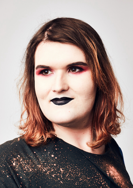 Headshot of white woman looking off camera and smiling. She has mid length red-brown hair and wears black lipstick, red eyeshadow, and wears a sparkly black dress.